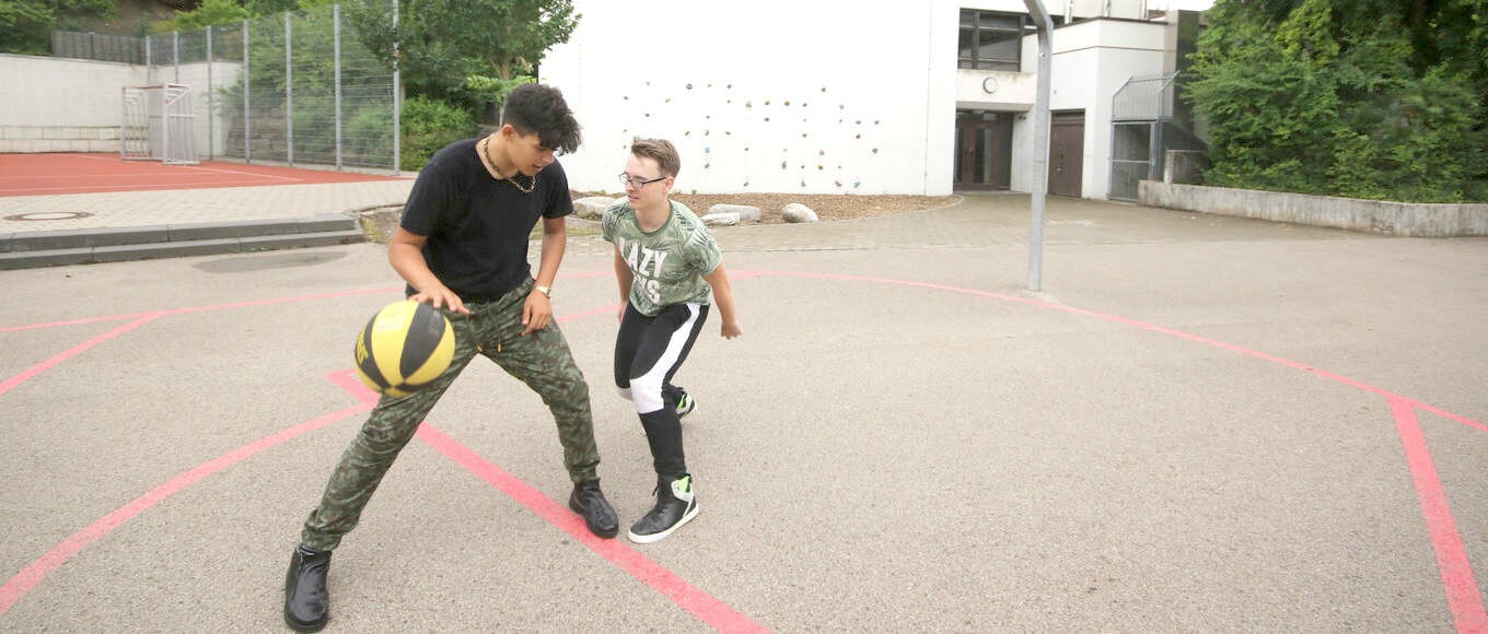 Zwei Jugendliche spielen Basketball auf einem großflächigen Sportplatz