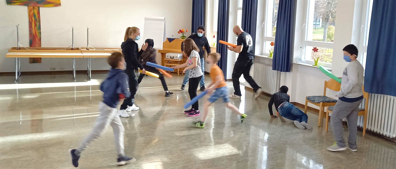 Eine Gruppe von Lehrern und Kindern spielen Poolnudeln-Fechten im Klassenraum